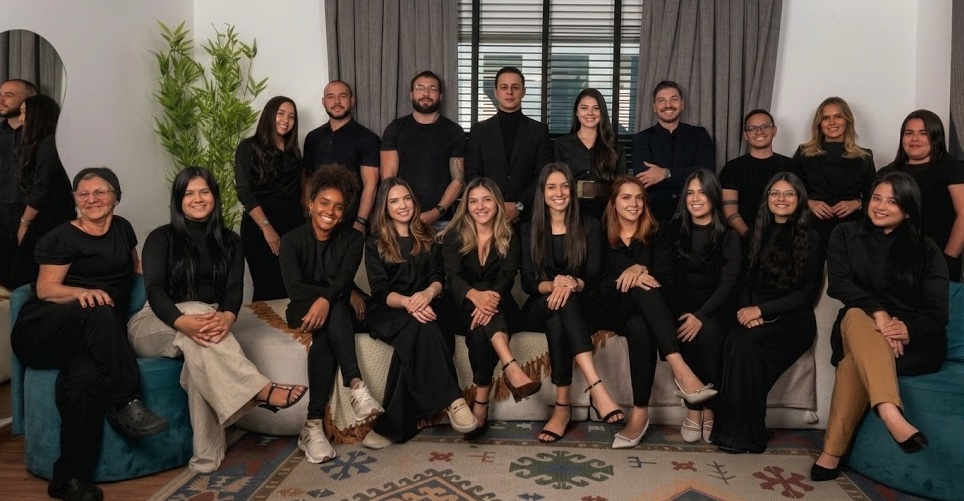 Equipe consolidada Dr. Lucas Moura: vinte profissionais com uniformes pretos reunidos em foto institucional, sentados em sofá e poltrona com tapete kilim de motivos geométricos em ambiente claro com cortinas e planta natural ao fundo.