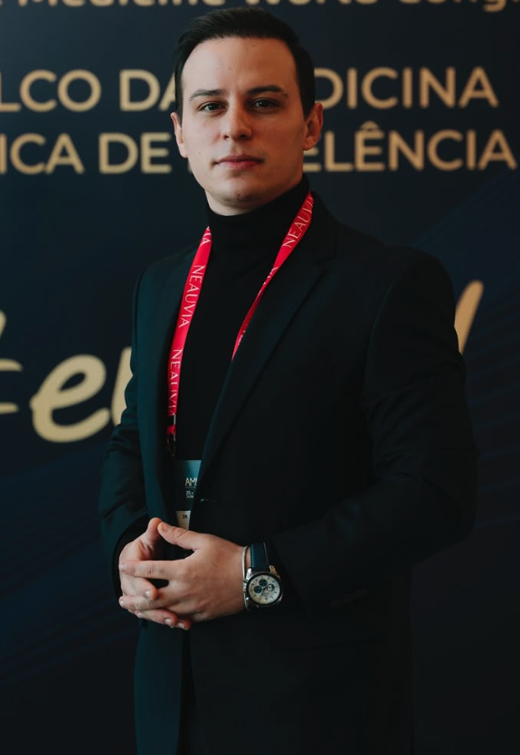 Retrato em pé do Dr. Lucas Moura em terno preto e gola alta, mãos sobrepostas em postura institucional, cordão e credencial de congresso médico internacional ao pescoço, expressão serena em ambiente de evento profissional.